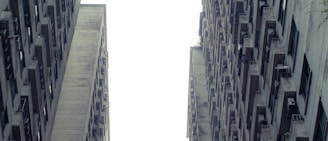 Two tall, aged concrete apartment buildings with numerous small balconies and windows stretch upwards, forming a narrow gap between them. The perspective creates a sense of height and verticality, with the sky visible as a bright contrast between the structures.