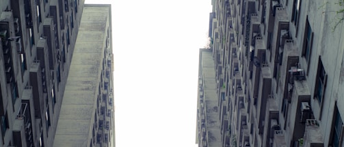 Two tall, aged concrete apartment buildings with numerous small balconies and windows stretch upwards, forming a narrow gap between them. The perspective creates a sense of height and verticality, with the sky visible as a bright contrast between the structures.