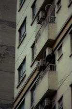 A close-up view of an older, worn-out residential building featuring several balconies with air conditioning units attached. The facade shows signs of decay, with peeled paint and a generally weathered appearance. Windows are aligned in vertical rows, and some balconies have rails.