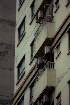 A close-up view of an older, worn-out residential building featuring several balconies with air conditioning units attached. The facade shows signs of decay, with peeled paint and a generally weathered appearance. Windows are aligned in vertical rows, and some balconies have rails.