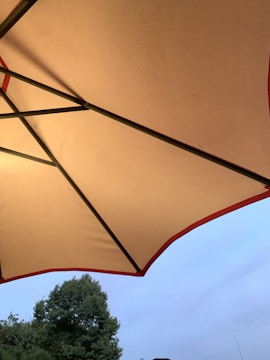 A large outdoor umbrella with a light beige fabric and a red edge is open, covering the view of the sky and a green tree in the background. The metal frame of the umbrella is visible and runs diagonally across the image.
