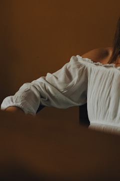 A close-up of a person's shoulder and arm in a white, off-shoulder blouse against a warm, brown background, focusing on the texture of the fabric.