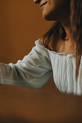 A close-up view of a person wearing a white, off-the-shoulder blouse. The lighting is warm and soft, highlighting the fabric's texture and creating a relaxed atmosphere.