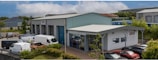 A commercial property featuring two buildings with metal roofs, situated in an industrial area. Several parked vehicles, including vans and cars, are seen near the buildings. The surroundings are well-maintained with greenery and clear weather with blue skies and clouds in the background.