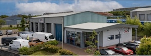 A commercial property featuring two buildings with metal roofs, situated in an industrial area. Several parked vehicles, including vans and cars, are seen near the buildings. The surroundings are well-maintained with greenery and clear weather with blue skies and clouds in the background.