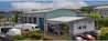 A commercial property featuring two buildings with metal roofs, situated in an industrial area. Several parked vehicles, including vans and cars, are seen near the buildings. The surroundings are well-maintained with greenery and clear weather with blue skies and clouds in the background.