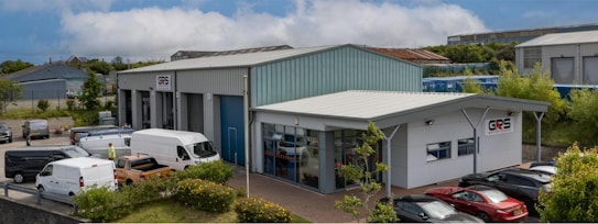 A commercial property featuring two buildings with metal roofs, situated in an industrial area. Several parked vehicles, including vans and cars, are seen near the buildings. The surroundings are well-maintained with greenery and clear weather with blue skies and clouds in the background.