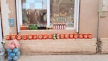 A storefront display showcases various beverages and snack items. Several bottles of colorful drinks, including green glass bottles, are lined up on the windowsill. Below the window, a neat row of red snack bags is arranged on a brick ledge. To the left, a stack of blue and pink striped balls adds a playful touch. The background reveals a reflection of a street scene in the glass window.