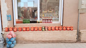 A storefront display showcases various beverages and snack items. Several bottles of colorful drinks, including green glass bottles, are lined up on the windowsill. Below the window, a neat row of red snack bags is arranged on a brick ledge. To the left, a stack of blue and pink striped balls adds a playful touch. The background reveals a reflection of a street scene in the glass window.