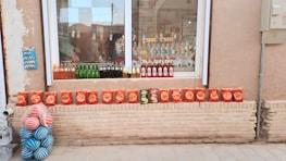 A storefront display showcases various beverages and snack items. Several bottles of colorful drinks, including green glass bottles, are lined up on the windowsill. Below the window, a neat row of red snack bags is arranged on a brick ledge. To the left, a stack of blue and pink striped balls adds a playful touch. The background reveals a reflection of a street scene in the glass window.