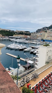 A scenic view of a marina filled with luxurious yachts and boats. Nearby, a row of bicycles is parked along a terrace overlooking the harbor. The background features a hillside with densely built structures and greenery, while some taller buildings are visible near the waterfront.