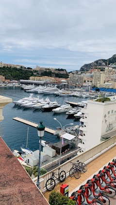 A scenic view of a marina filled with luxurious yachts and boats. Nearby, a row of bicycles is parked along a terrace overlooking the harbor. The background features a hillside with densely built structures and greenery, while some taller buildings are visible near the waterfront.