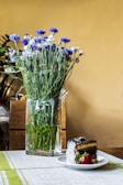 A bouquet of blue and white wildflowers is arranged in a tall, clear glass vase placed on a table. Beside the vase, a slice of cake with chocolate icing and coconut flakes is served on a white plate, accompanied by a fresh strawberry. The table is covered with a light-colored runner, and the background has a warm yellow hue.