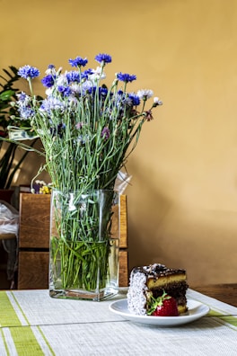 A bouquet of blue and white wildflowers is arranged in a tall, clear glass vase placed on a table. Beside the vase, a slice of cake with chocolate icing and coconut flakes is served on a white plate, accompanied by a fresh strawberry. The table is covered with a light-colored runner, and the background has a warm yellow hue.
