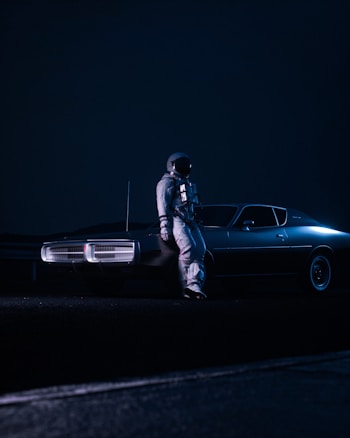 An astronaut in a space suit stands next to a vintage car in a dimly lit, surreal nighttime setting. The scene conveys a futuristic or alternate reality atmosphere with a dark blue color palette dominating the environment. The astronaut appears contemplative, leaning against the vehicle under the expansive dark sky.