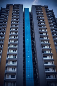 A tall apartment building with a modern architectural design features a distinctive blue glass facade in the center and flanking concrete walls with balconies and windows.
