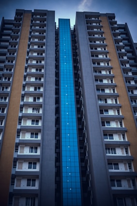 A tall apartment building with a modern architectural design features a distinctive blue glass facade in the center and flanking concrete walls with balconies and windows.