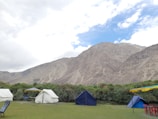 A campsite is set against a backdrop of rugged mountains and a partly cloudy sky. There are several tents pitched on a grassy area, with some chairs and a table nearby. The landscape is lush with green shrubs and the tents are mainly white and blue.
