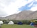 A campsite is set against a backdrop of rugged mountains and a partly cloudy sky. There are several tents pitched on a grassy area, with some chairs and a table nearby. The landscape is lush with green shrubs and the tents are mainly white and blue.