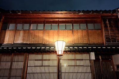 A traditional wooden building with a soft, warm glow illuminating from a central lantern. The structure features wooden panels and rectangular windows, radiating an atmospheric, rustic charm.