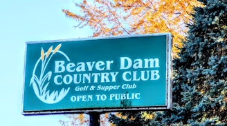 A signboard for Beaver Dam Country Club, featuring the text 'Golf & Supper Club' and 'Open to Public.' The sign is set amidst natural surroundings with autumn-colored leaves and evergreen trees.