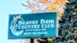 A signboard for Beaver Dam Country Club, featuring the text 'Golf & Supper Club' and 'Open to Public.' The sign is set amidst natural surroundings with autumn-colored leaves and evergreen trees.
