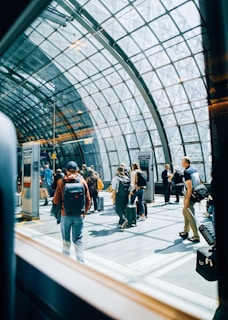 A group of people with luggage are moving through a modern, glass-roofed terminal. Natural light floods the space, creating reflections on the floor. The architecture features a grid-like metal framework supporting the glass ceiling, giving the area an open and airy feel.