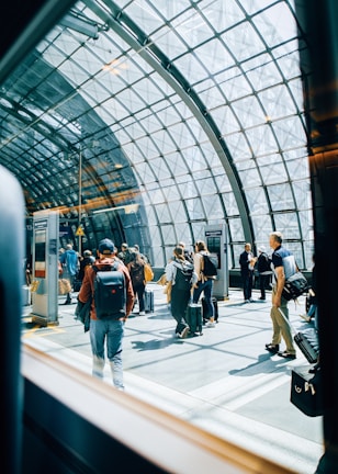 A group of people with luggage are moving through a modern, glass-roofed terminal. Natural light floods the space, creating reflections on the floor. The architecture features a grid-like metal framework supporting the glass ceiling, giving the area an open and airy feel.