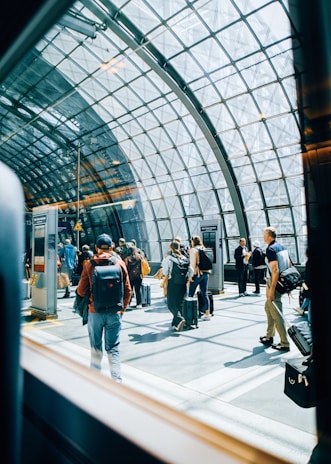 A group of people with luggage are moving through a modern, glass-roofed terminal. Natural light floods the space, creating reflections on the floor. The architecture features a grid-like metal framework supporting the glass ceiling, giving the area an open and airy feel.