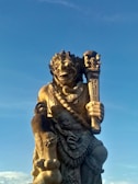 A large stone statue of a mythological figure with intricate carvings. The figure appears to hold a staff or club and is adorned with detailed jewelry and clothing. The background is a clear blue sky, indicating it is outdoors and possibly part of a cultural or religious site.