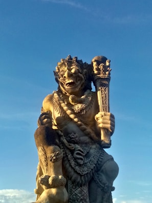 A large stone statue of a mythological figure with intricate carvings. The figure appears to hold a staff or club and is adorned with detailed jewelry and clothing. The background is a clear blue sky, indicating it is outdoors and possibly part of a cultural or religious site.