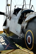 Close-up view of a vintage military aircraft cockpit, featuring a circular roundel insignia on the side. The cockpit is partially open, revealing control gauges and instruments. Historical aviation elements like wires and leather straps are visible, with the aircraft painted in muted military colors.