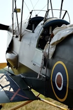 Close-up view of a vintage military aircraft cockpit, featuring a circular roundel insignia on the side. The cockpit is partially open, revealing control gauges and instruments. Historical aviation elements like wires and leather straps are visible, with the aircraft painted in muted military colors.