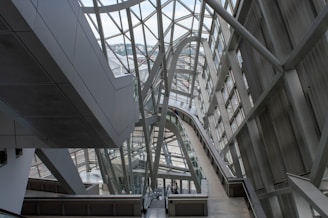 A modern architectural interior featuring large metal and glass structures, creating an intricate network of beams and walkways. The design is sleek and futuristic, with ample natural light pouring in through the windows lining the walls and ceiling.