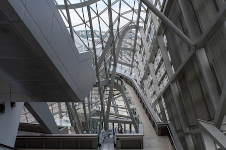 A modern architectural interior featuring large metal and glass structures, creating an intricate network of beams and walkways. The design is sleek and futuristic, with ample natural light pouring in through the windows lining the walls and ceiling.