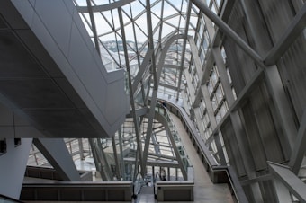 A modern architectural interior featuring large metal and glass structures, creating an intricate network of beams and walkways. The design is sleek and futuristic, with ample natural light pouring in through the windows lining the walls and ceiling.
