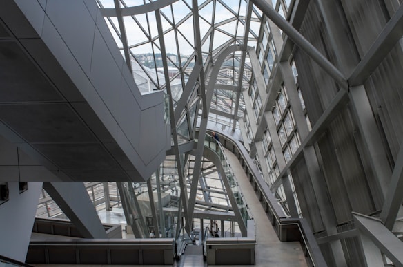 A modern architectural interior featuring large metal and glass structures, creating an intricate network of beams and walkways. The design is sleek and futuristic, with ample natural light pouring in through the windows lining the walls and ceiling.