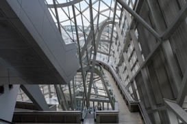 A modern architectural interior featuring large metal and glass structures, creating an intricate network of beams and walkways. The design is sleek and futuristic, with ample natural light pouring in through the windows lining the walls and ceiling.