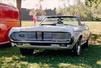 A classic white convertible car with a Canadian flag on display in a grassy area. The car is parked alongside a red vehicle. Trees and a building are visible in the background.