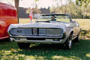 A classic white convertible car with a Canadian flag on display in a grassy area. The car is parked alongside a red vehicle. Trees and a building are visible in the background.