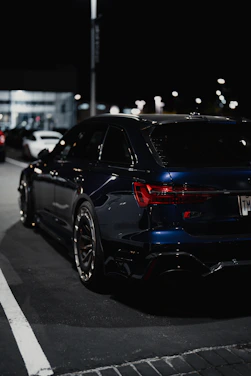 A modern, dark-themed photo of a shiny black car parked under city lights at night.