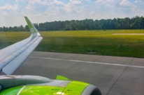 An airplane wing with a vibrant green engine is visible as the aircraft taxis on a runway. The background features a grassy field and a line of trees under a partly cloudy sky.