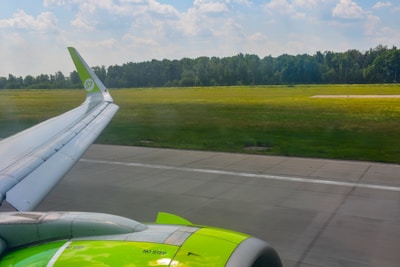 An airplane wing with a vibrant green engine is visible as the aircraft taxis on a runway. The background features a grassy field and a line of trees under a partly cloudy sky.