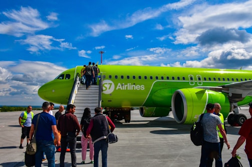 A vibrant green airplane with passengers boarding via an external staircase on a sunny day. The scene includes a clear blue sky with some clouds overhead, and several people, some carrying bags, are visible either entering the aircraft or standing near it.