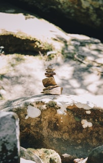 A small stack of balanced rocks on a larger moss-covered boulder, surrounded by dappled sunlight and shade. The rocks are arranged in a vertical alignment, showcasing an example of natural balance and tranquility.