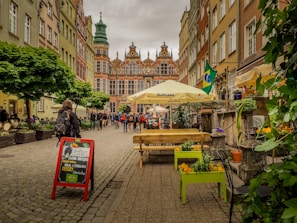 A cobblestone street lined with colorful historic buildings and a prominent building featuring intricate architecture is visible at the end. People are walking along the street, and a restaurant sign promotes Brazilian cuisine with flags and umbrellas adding vibrant touches. Green plants and flowers are arranged in planters along the pathway.