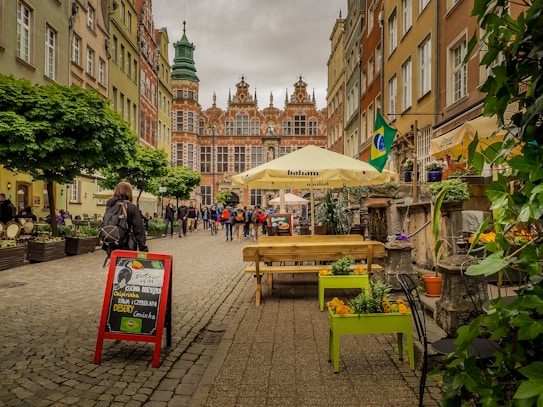 A cobblestone street lined with colorful historic buildings and a prominent building featuring intricate architecture is visible at the end. People are walking along the street, and a restaurant sign promotes Brazilian cuisine with flags and umbrellas adding vibrant touches. Green plants and flowers are arranged in planters along the pathway.