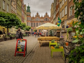 A cobblestone street lined with colorful historic buildings and a prominent building featuring intricate architecture is visible at the end. People are walking along the street, and a restaurant sign promotes Brazilian cuisine with flags and umbrellas adding vibrant touches. Green plants and flowers are arranged in planters along the pathway.