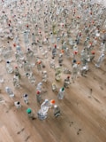 A large collection of plastic bottles of various shapes and sizes spread across a wooden floor. Some bottles are clear while others contain liquids of different colors. Many have colorful caps in shades like red, blue, green, and orange.