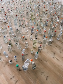 A large collection of plastic bottles of various shapes and sizes spread across a wooden floor. Some bottles are clear while others contain liquids of different colors. Many have colorful caps in shades like red, blue, green, and orange.
